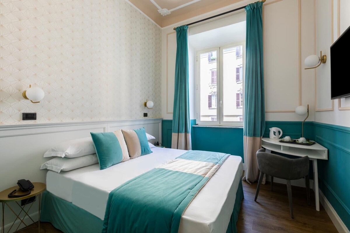 Habitación de hotel con cama, escritorio, silla, ventana con cortinas y televisión montada en la pared.