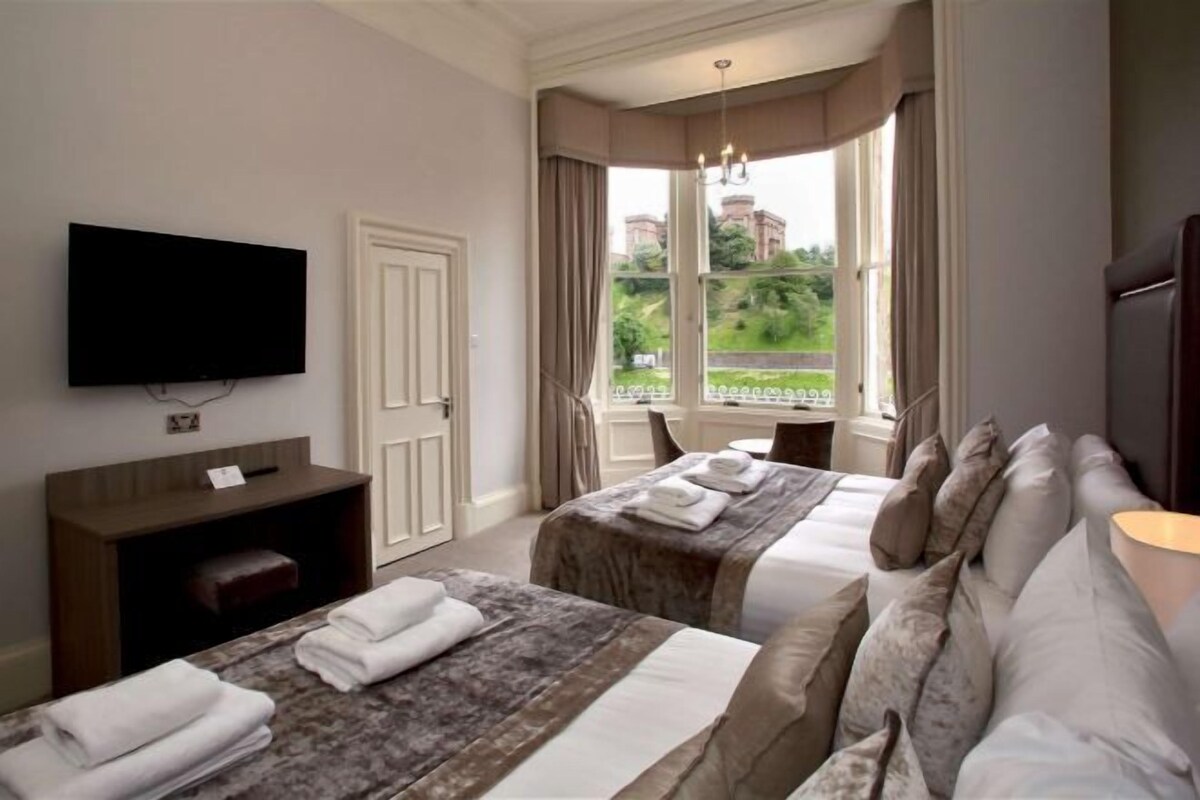 A hotel room with two beds, a TV, a desk, and a window with a view of buildings and greenery.