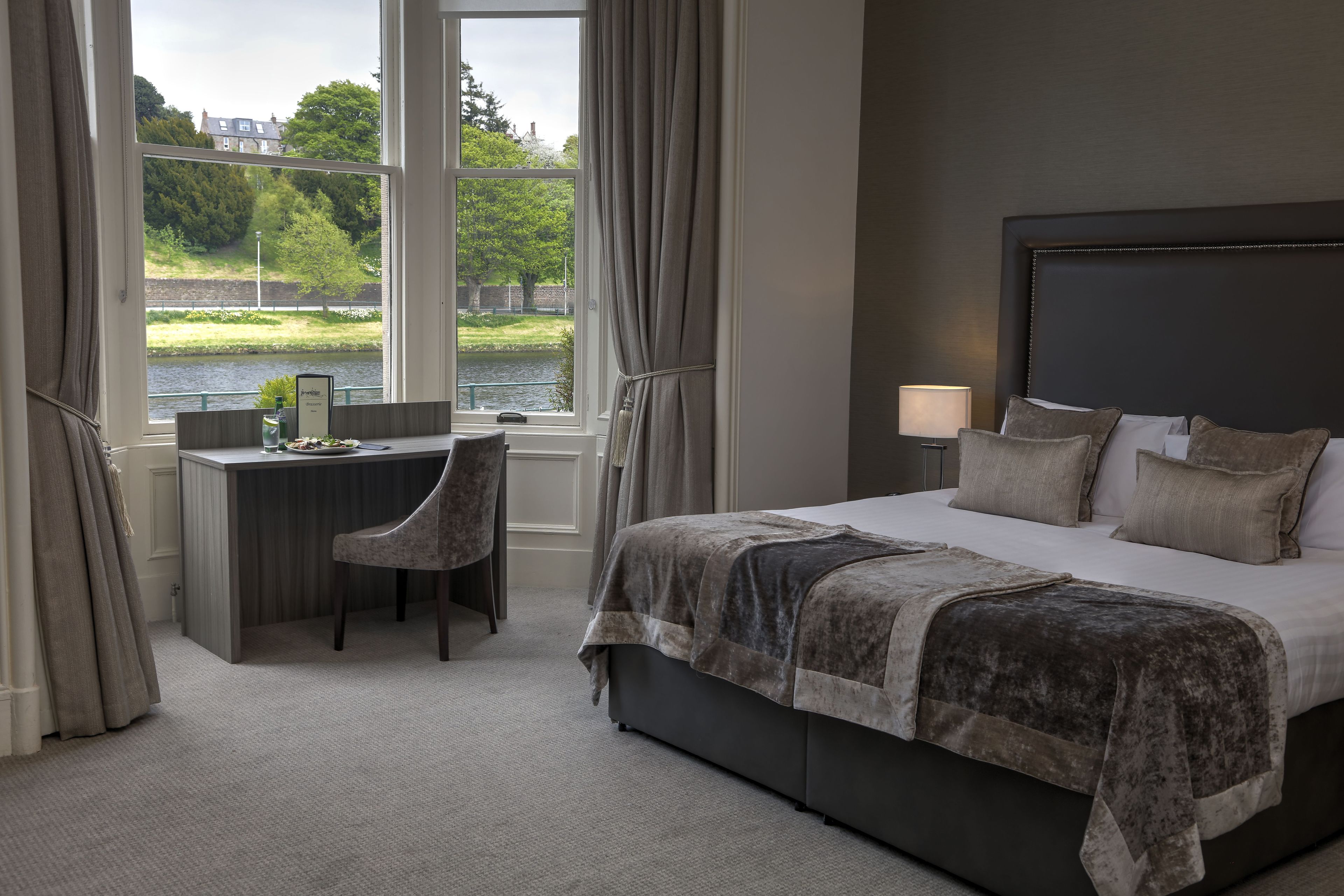 A bedroom with a large bed, a desk, and a chair. There is a window with a view of a river and trees.