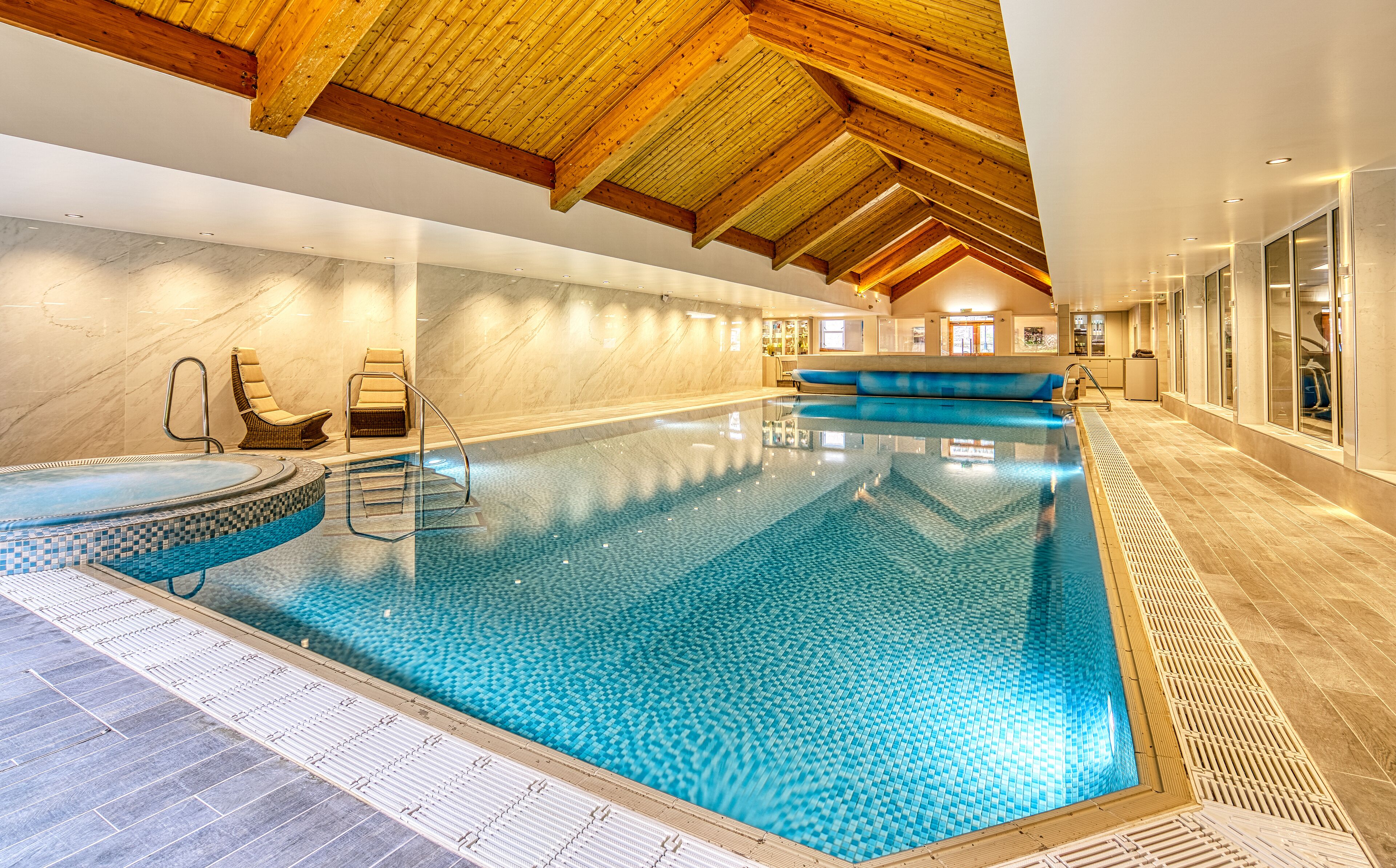 An indoor swimming pool with a tiled floor, wooden ceiling beams, and a seating area with a chair and a bench.