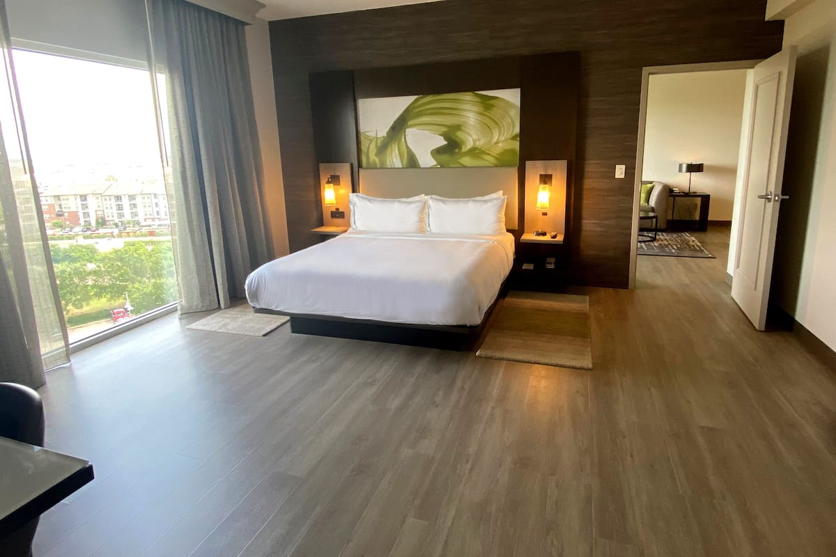 Ein modernes Hotelzimmer mit einem großen Bett, zwei Nachttischen mit Lampen sowie Blick auf Grünflächen und einer Tür zu einem weiteren Raum.