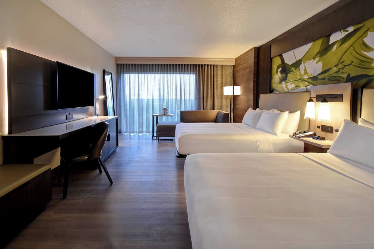 Ein Hotelzimmer mit zwei Betten, einem Schreibtisch mit Stuhl, einem Fernseher, einem kleinen Tisch und einem Fenster mit Vorhängen.
