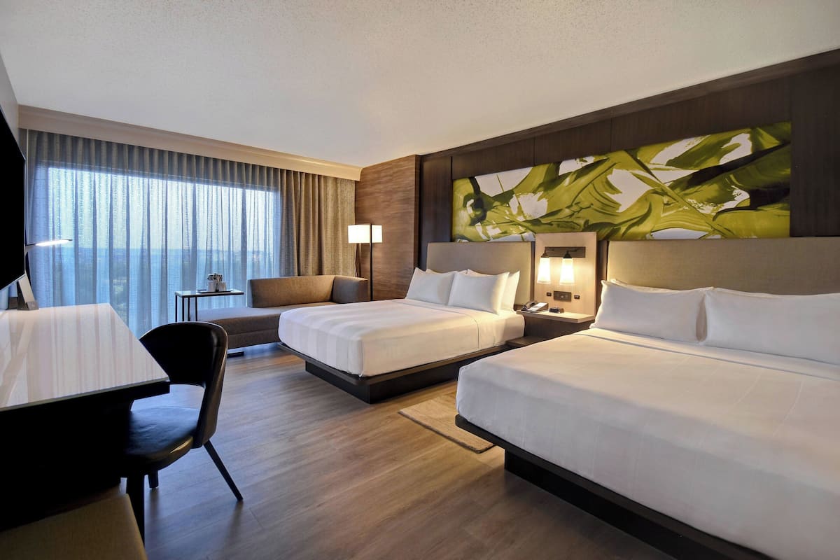 Ein Hotelzimmer mit zwei Betten, einem Schreibtisch, einem Stuhl, einer Lampe und einem großen abstrakten Gemälde an der Wand.