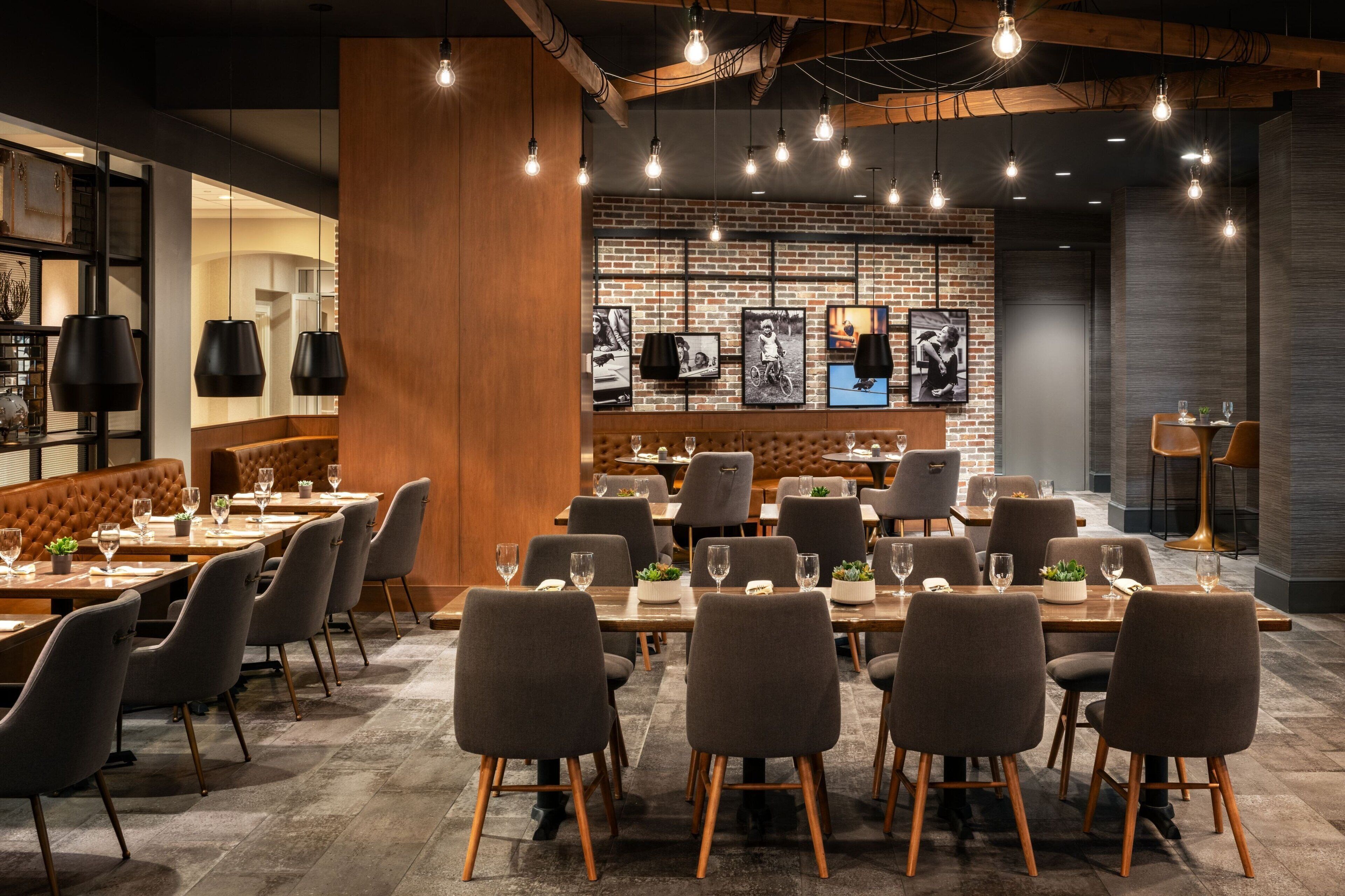 A modern restaurant with multiple dining tables and chairs, a brick wall with framed pictures, and hanging light fixtures.