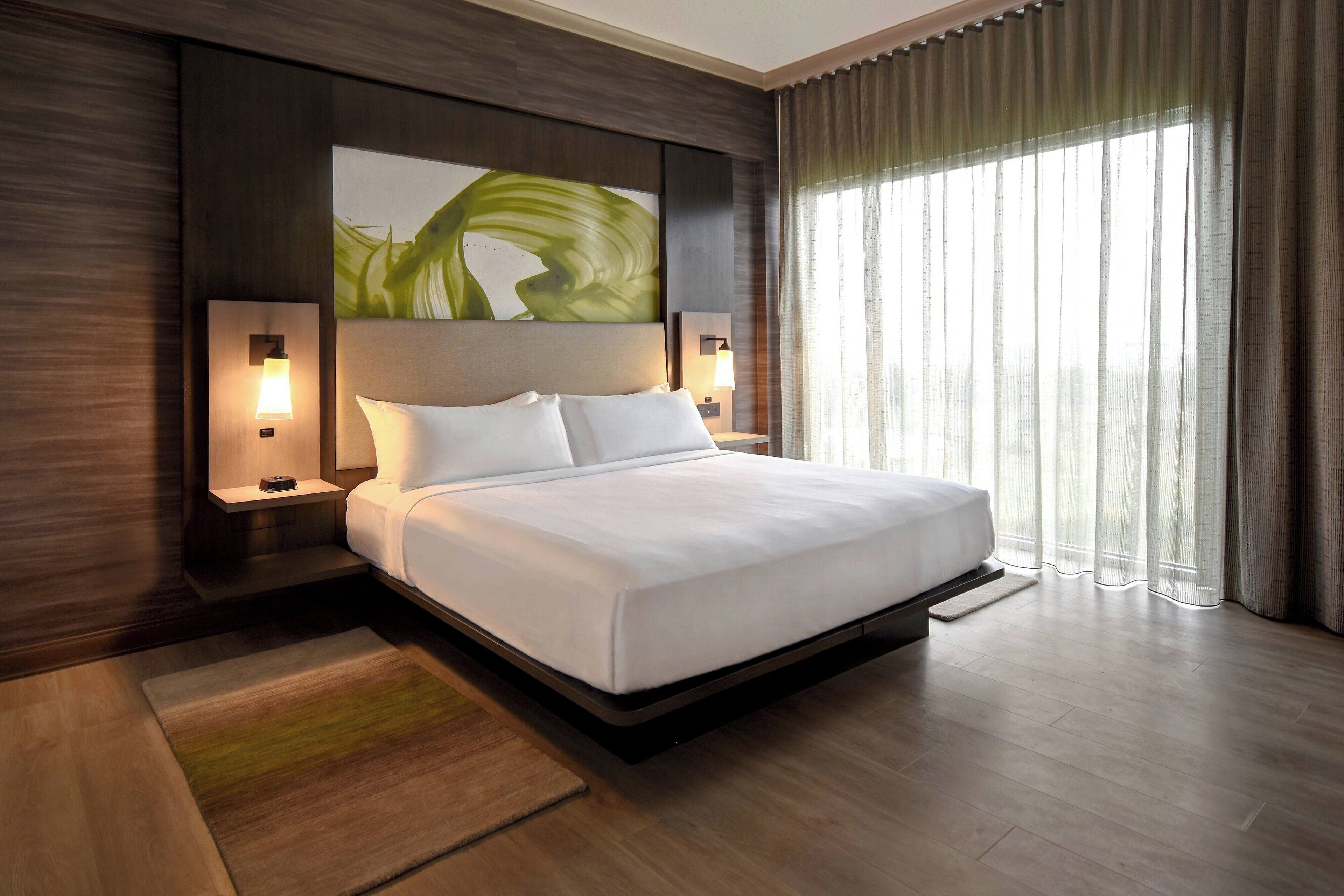 Un hotel moderno con una cama grande, dos mesitas de noche con lámparas, un cuadro abstracto en la pared y vistas de la ciudad a través de cortinas translúcidas.