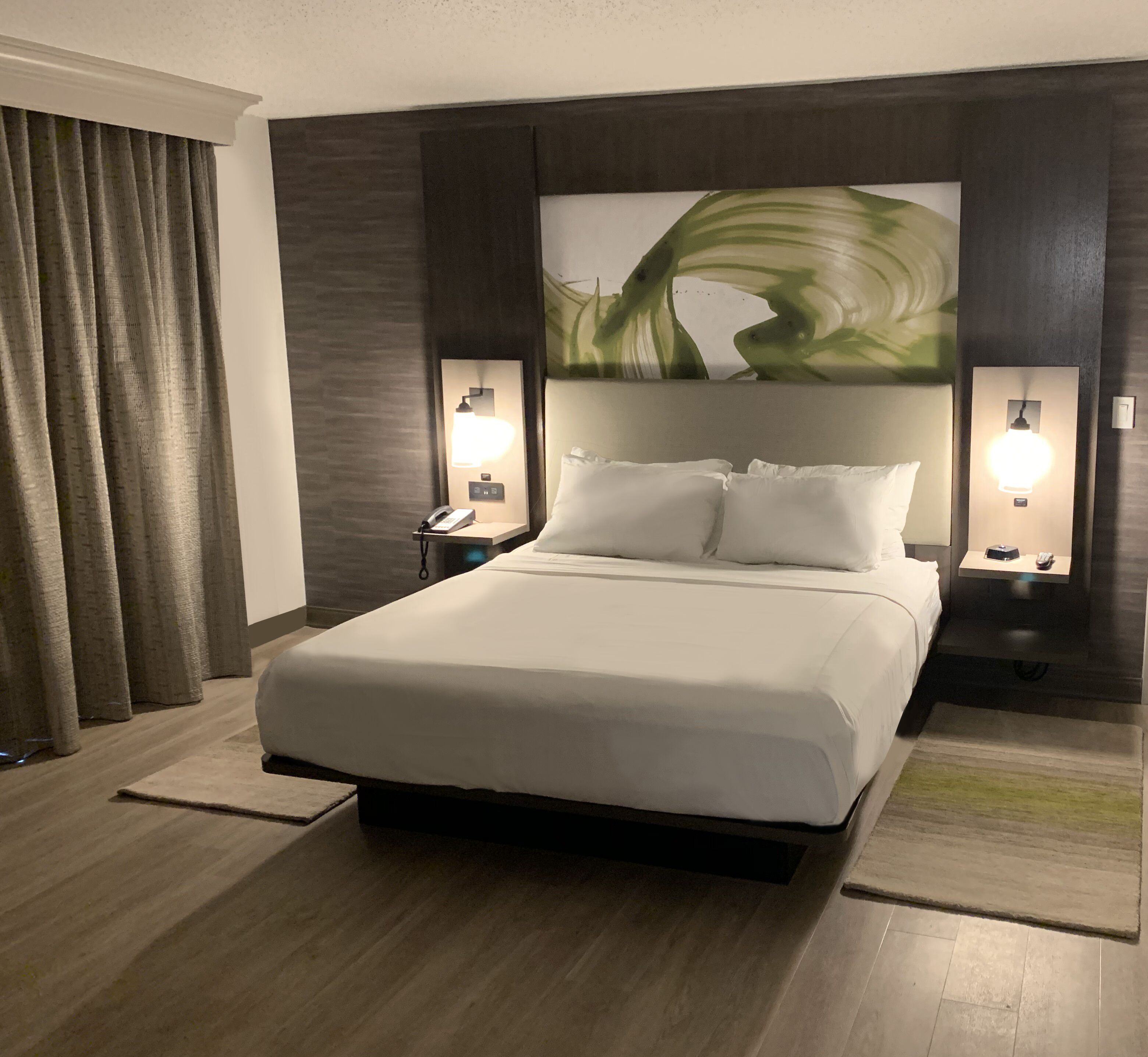 A hotel room with a large bed, two bedside tables with lamps, a large abstract painting on the wall, and a window with curtains.