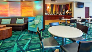 Un hall d’hôtel doté d’un espace salon comprenant un canapé, des fauteuils et une table ronde. On aperçoit à l’arrière-plan un bar avec un comptoir et divers éléments.