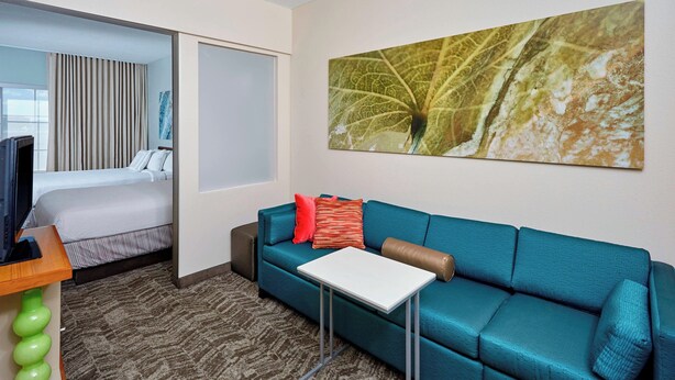 Une chambre d’hôtel avec un lit, un canapé de couleur turquoise, une petite table et une grande œuvre d’art représentant des motifs de feuilles accrochée au mur.