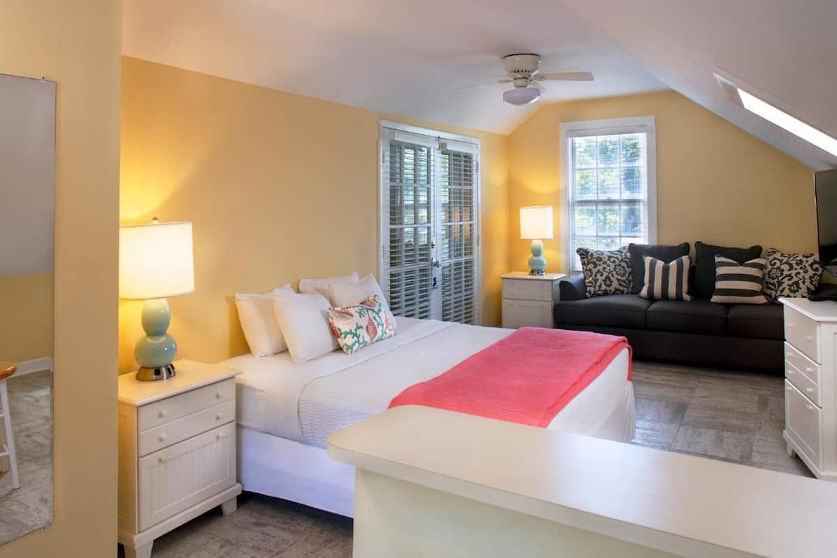 Un dormitorio con cama, mesitas de noche, cómoda, ventana con persianas, ventilador de techo y sofá.