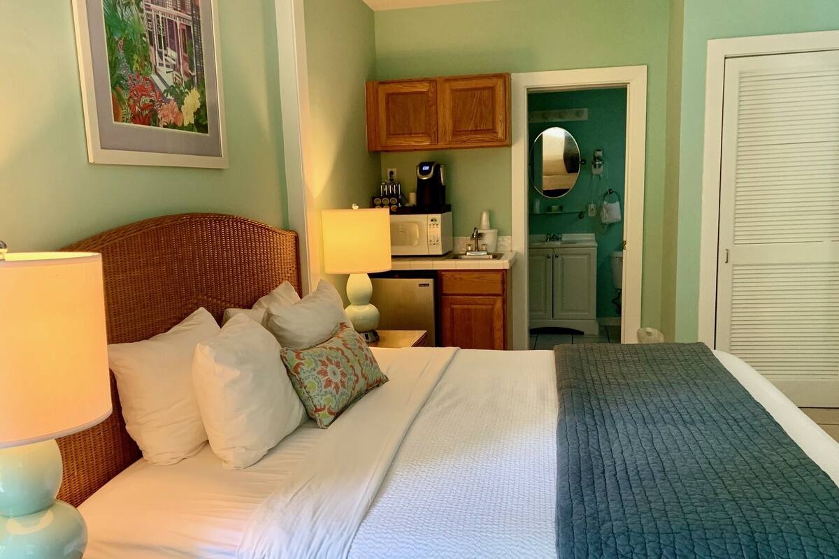 Una habitación de hotel con cama, lámpara de noche, mesita, microondas, lavamanos y espejo.
