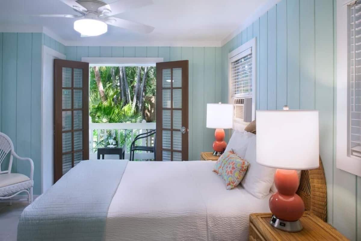 Un dormitorio con cama, dos lámparas de noche, ventilador de techo y vistas a un balcón con plantas.