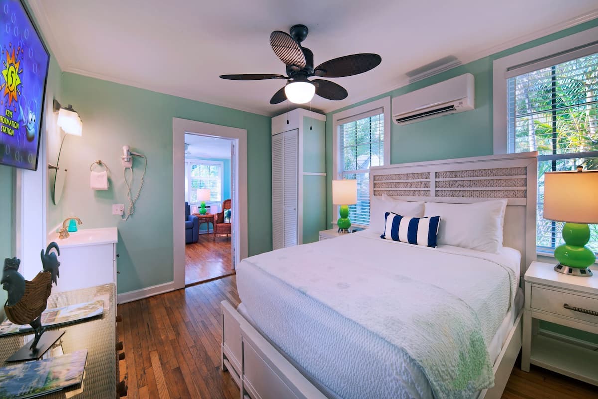 Un dormitorio con cama, ventilador de techo, mesita de noche, lámpara y ventana.