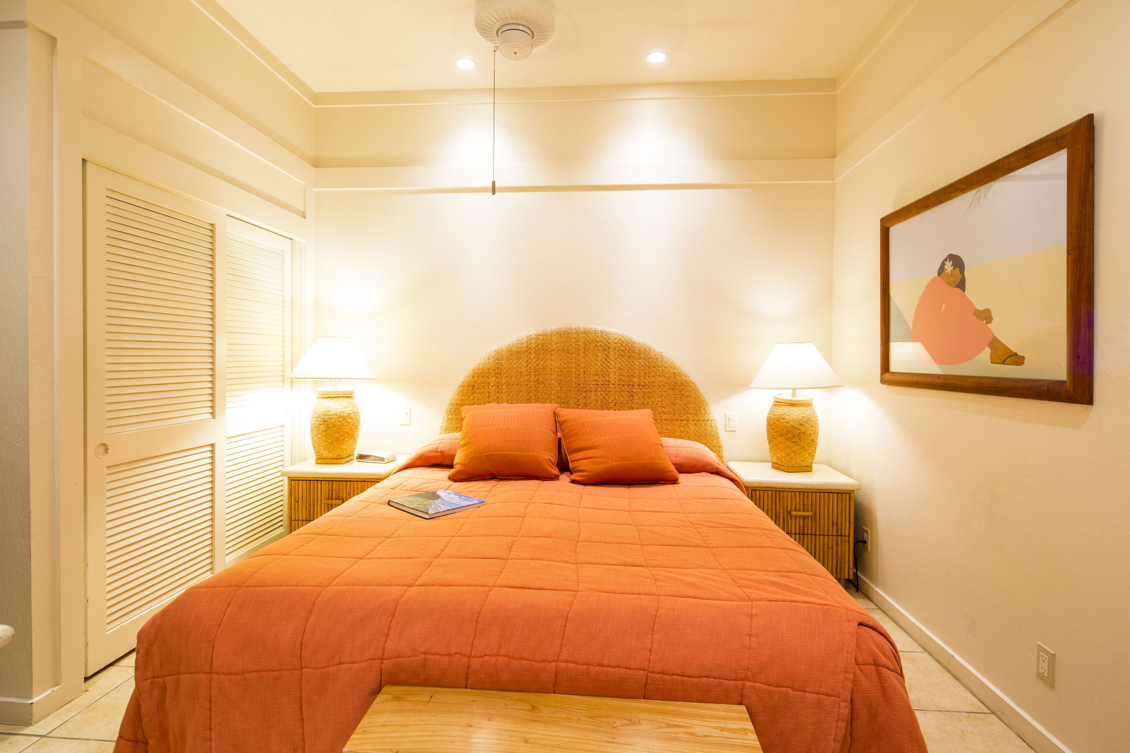 Una cama bien hecha con una colcha y almohadas naranjas, dos lámparas de noche y un cuadro en la pared.