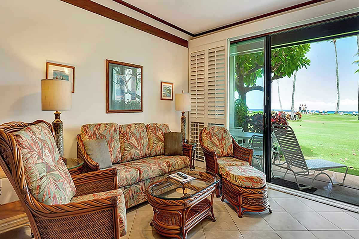 A living room with a sofa, chairs, a coffee table, and a large glass door leading to an outdoor area with a lawn and palm trees.