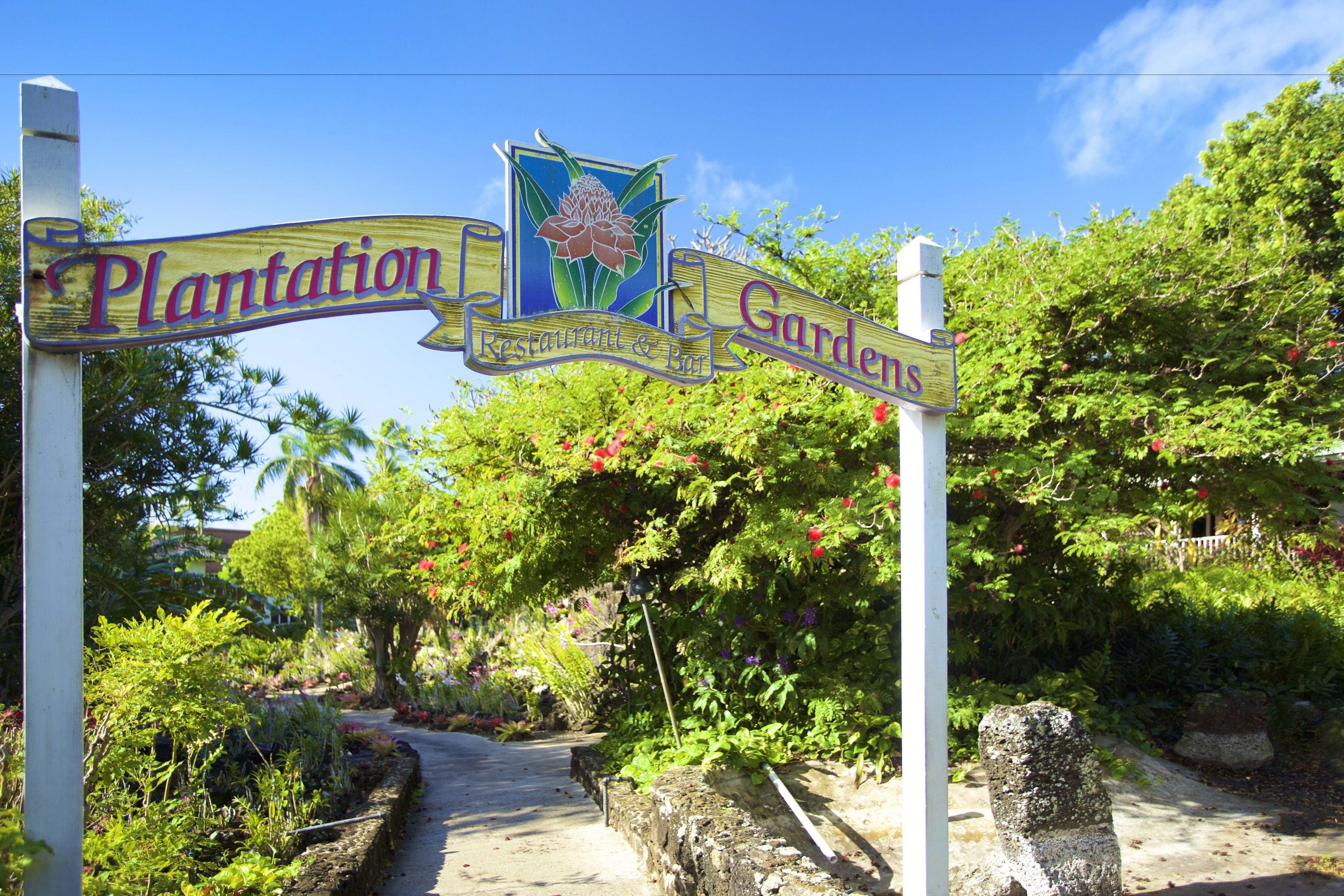 A sign for Plantation Gardens with a floral design and a pathway leading through lush greenery.