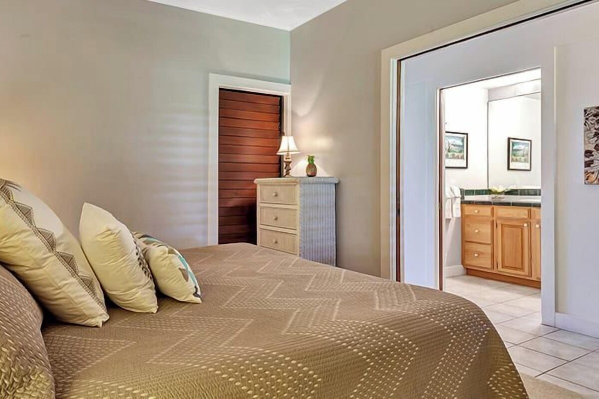 Un dormitorio con cama, almohadas, una mesita de noche, una lámpara, un aparador y una puerta que da a un baño.