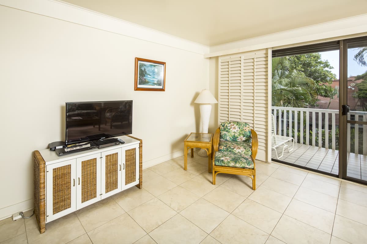 A living room with a TV on a white cabinet, a chair with a floral cushion, a small table with a lamp, and a balcony with a white railing.
