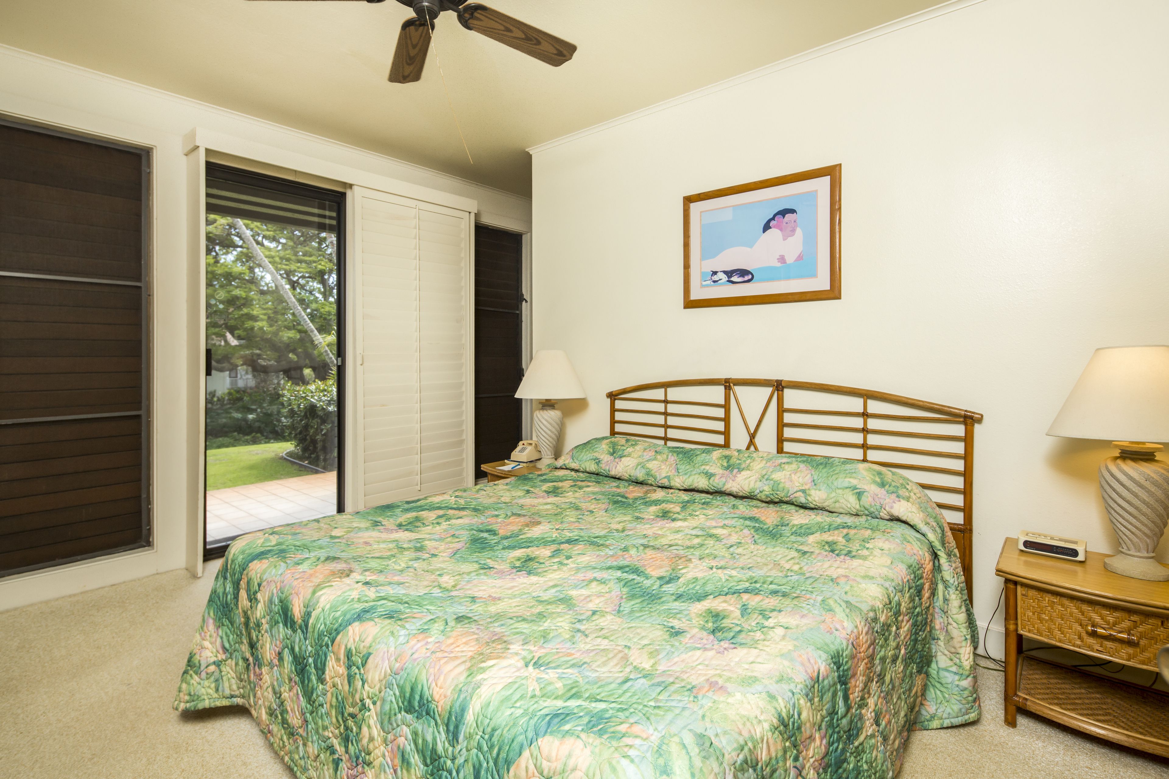 A bedroom with a bed, a nightstand, a lamp, a ceiling fan, a window with a view of the outdoors, and a framed picture on the wall.