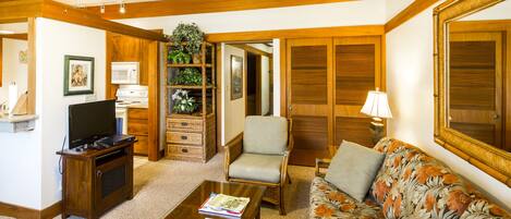 A living room with a wooden ceiling fan, a television on a wooden stand, a floral sofa, a chair, a coffee table with books, a lamp, a microwave, and a wicker basket with plants.