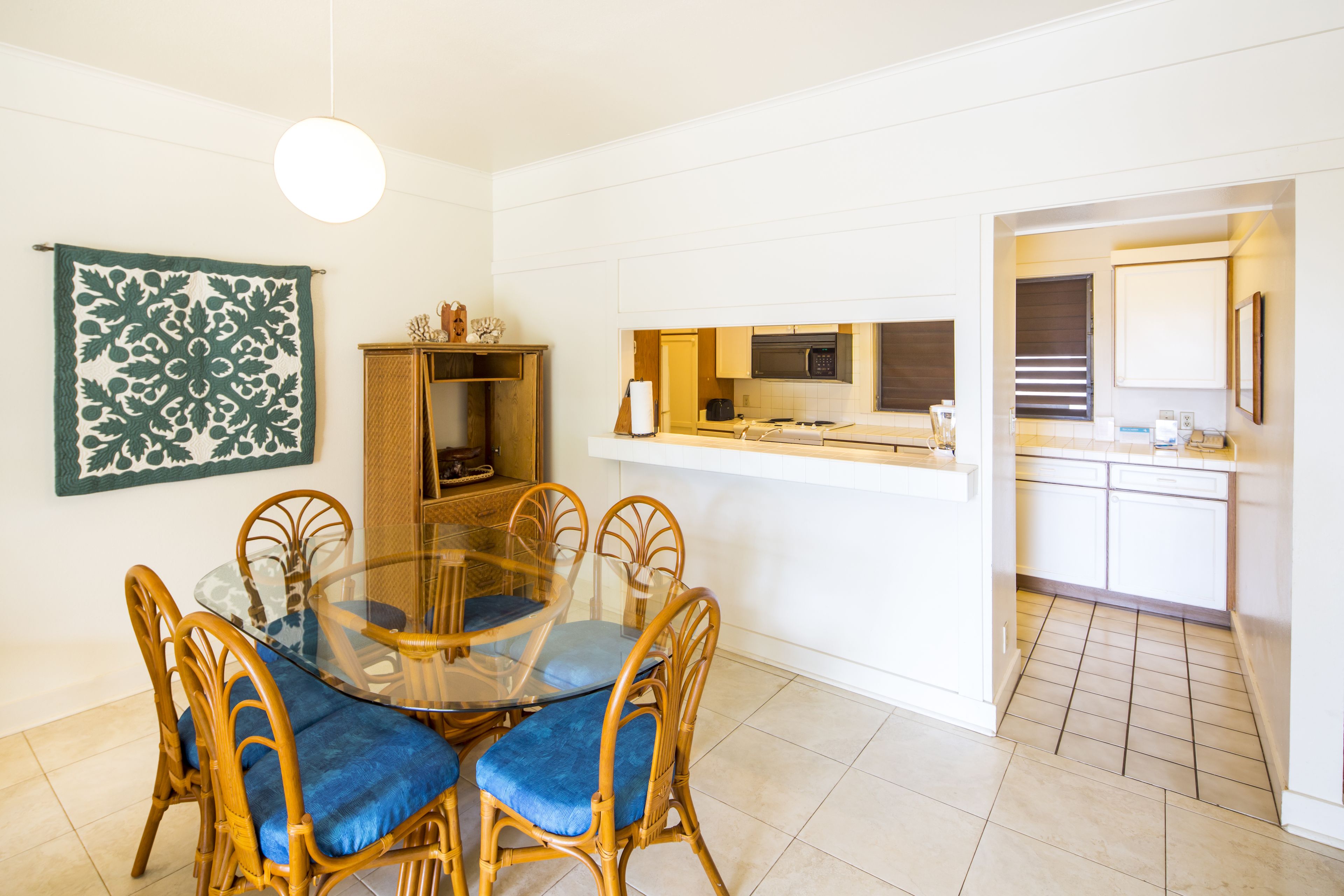 Un comedor con mesa de vidrio y sillas con acolchado azul, una cocina con armarios blancos y un tapiz de pared estampado.