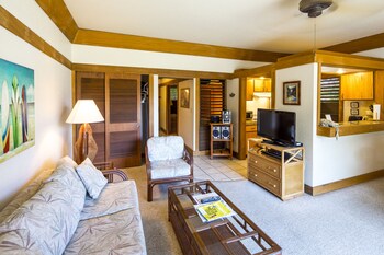 A dining area with wicker chairs and a glass table, a kitchen with modern appliances, and a painting of a landscape on the wall. at Castle Kiahuna Plantation & Beach Bungalows