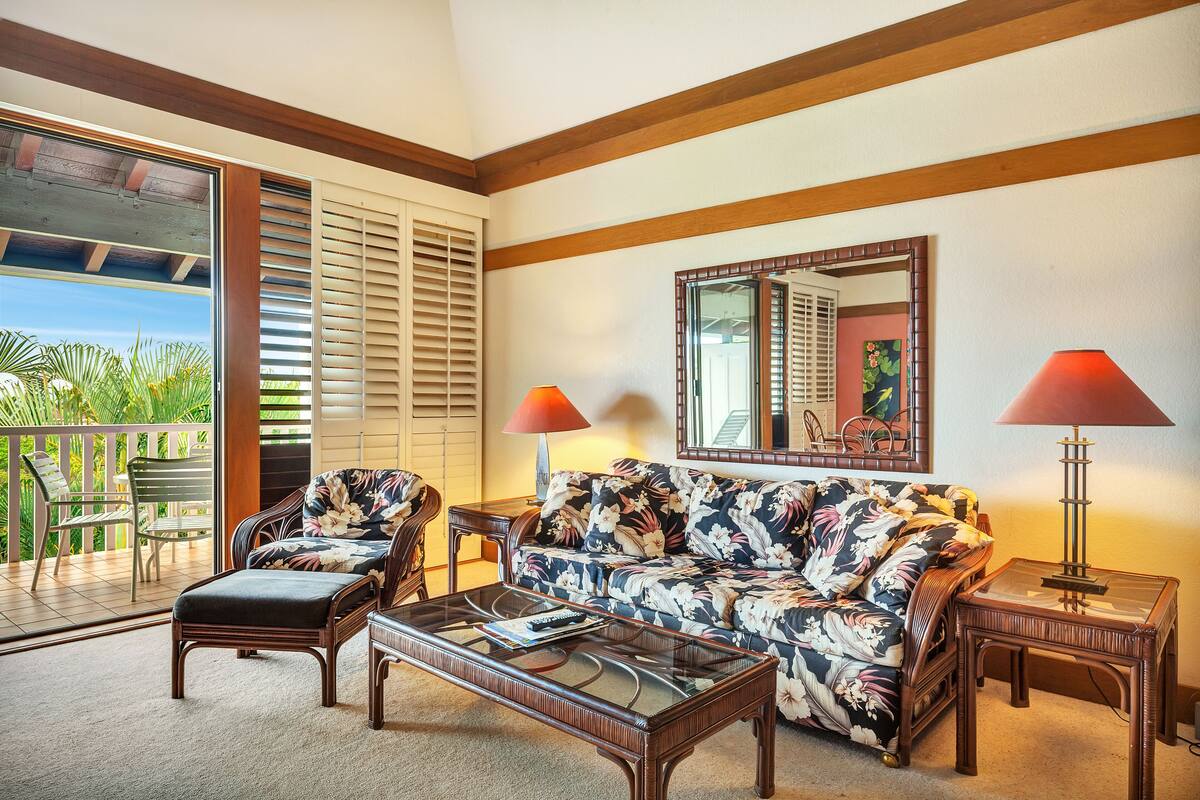 A living room with a sofa, two armchairs, a coffee table, and a glass-top side table. There is a large window with blinds, a balcony with chairs, and a painting on the wall.