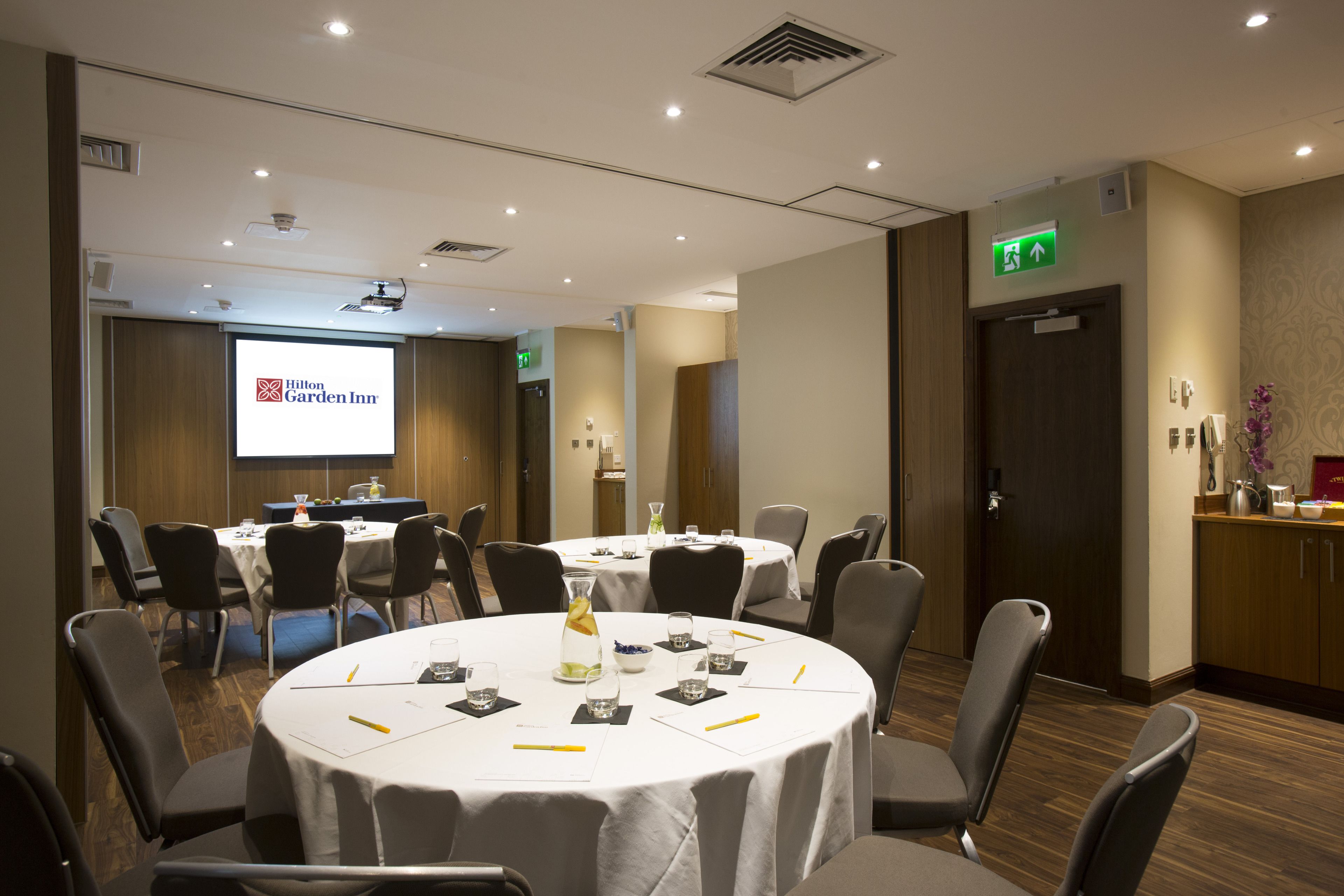 Une salle de conférence dotée d’un grand écran affichant le logo « Hilton Garden Inn », de tables rondes avec des chaises, et d’un espace de réception avec un comptoir et des plantes décoratives.