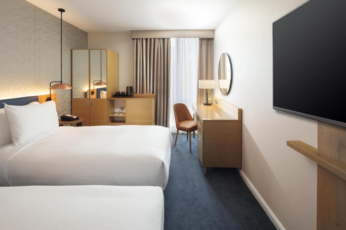 Een hotelkamer met twee bedden, een bureau, een stoel, een televisie, een spiegel en een raam met gordijnen.