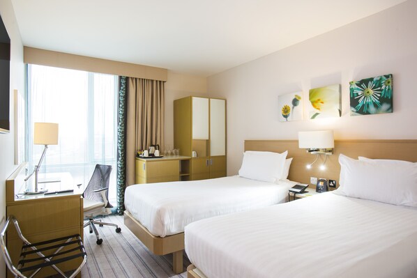 A hotel room with two beds, a desk, a chair, a desk lamp, a telephone, and artwork on the wall.