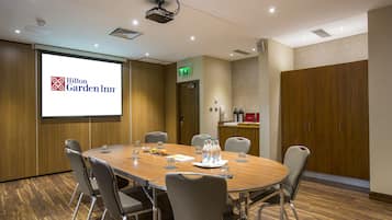 A conference room with a large screen displaying Hilton Garden Inn logo, a wooden table, and grey chairs.