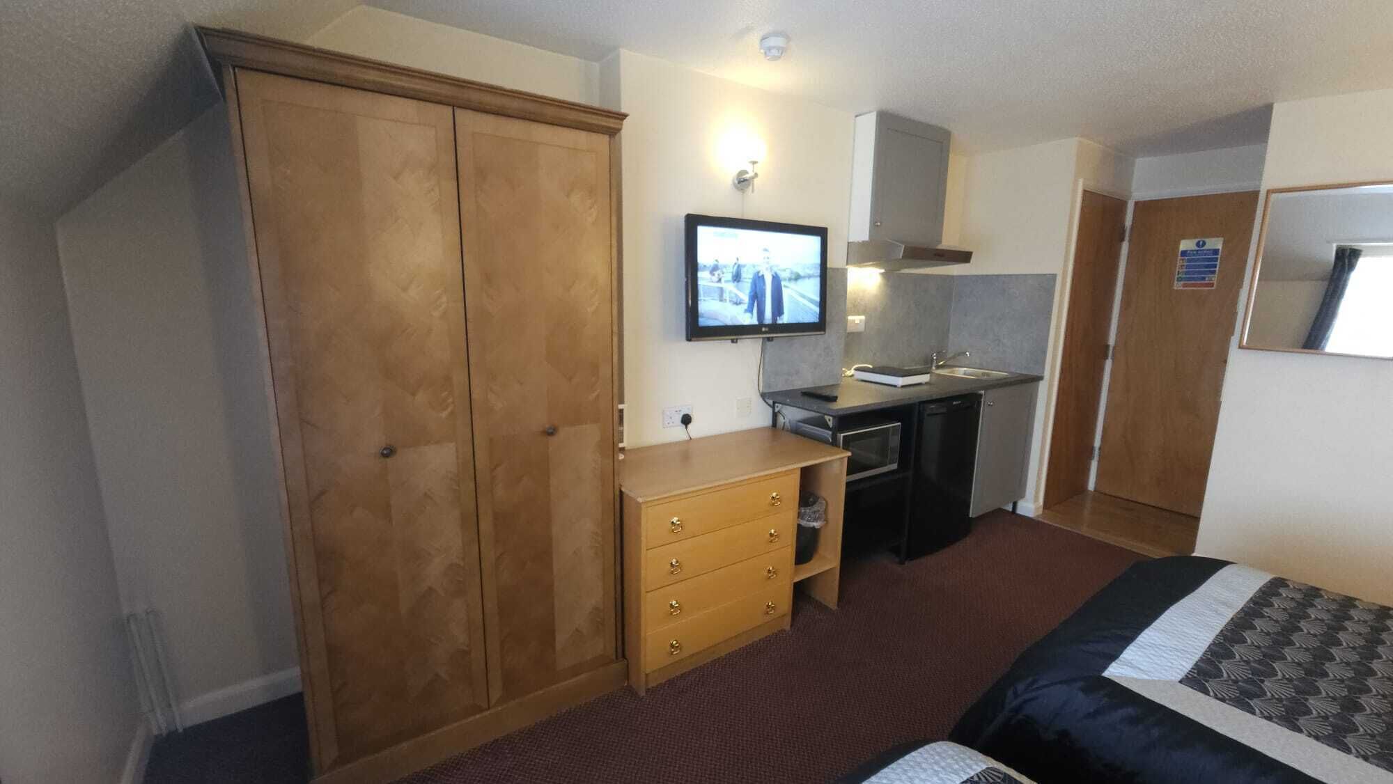 A hotel room with a wardrobe, a small kitchenette, a bed, and a television.