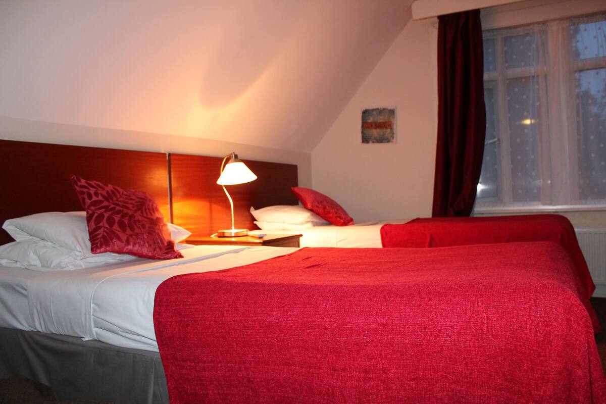 A hotel room with two beds, a nightstand with a lamp, and a window with curtains.