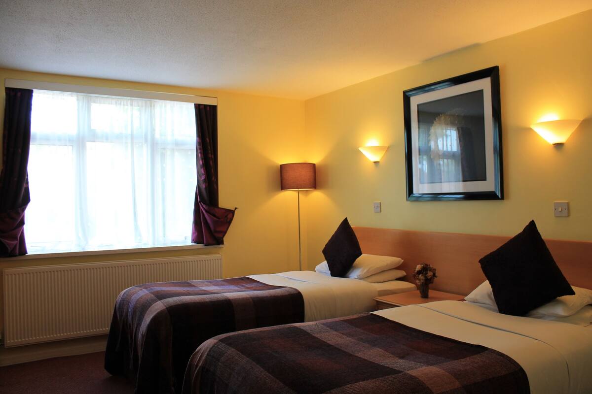 A hotel room with two beds, a large window with curtains, a standing lamp, and a framed picture on the wall.