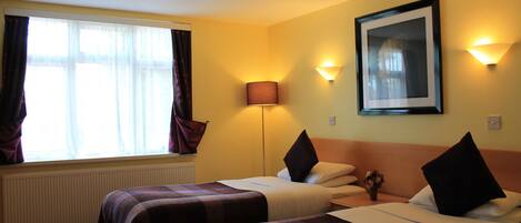 Een hotelkamer met twee bedden, een groot raam met gordijnen, een staande lamp en een lijst met een afbeelding aan de muur.