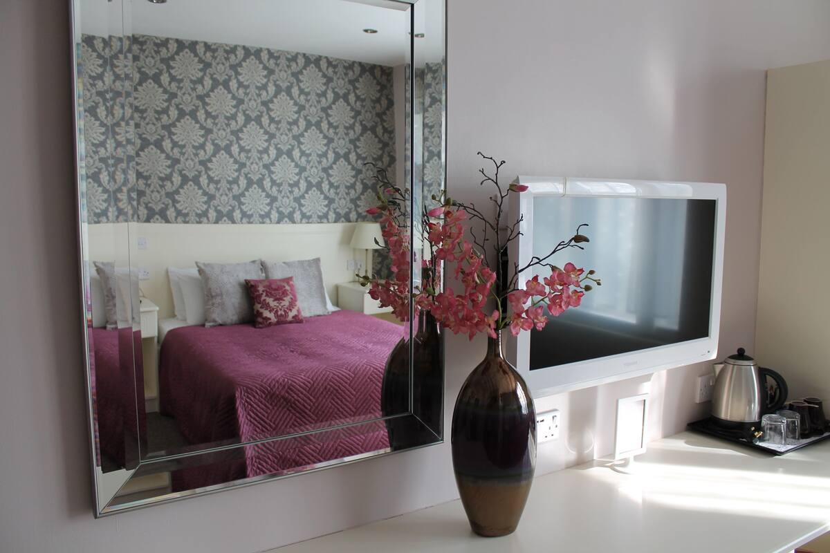 A bedroom with a large mirror, a bed with a pink bedspread, a vase with pink flowers, a flat-screen TV, and a coffee maker on the nightstand.