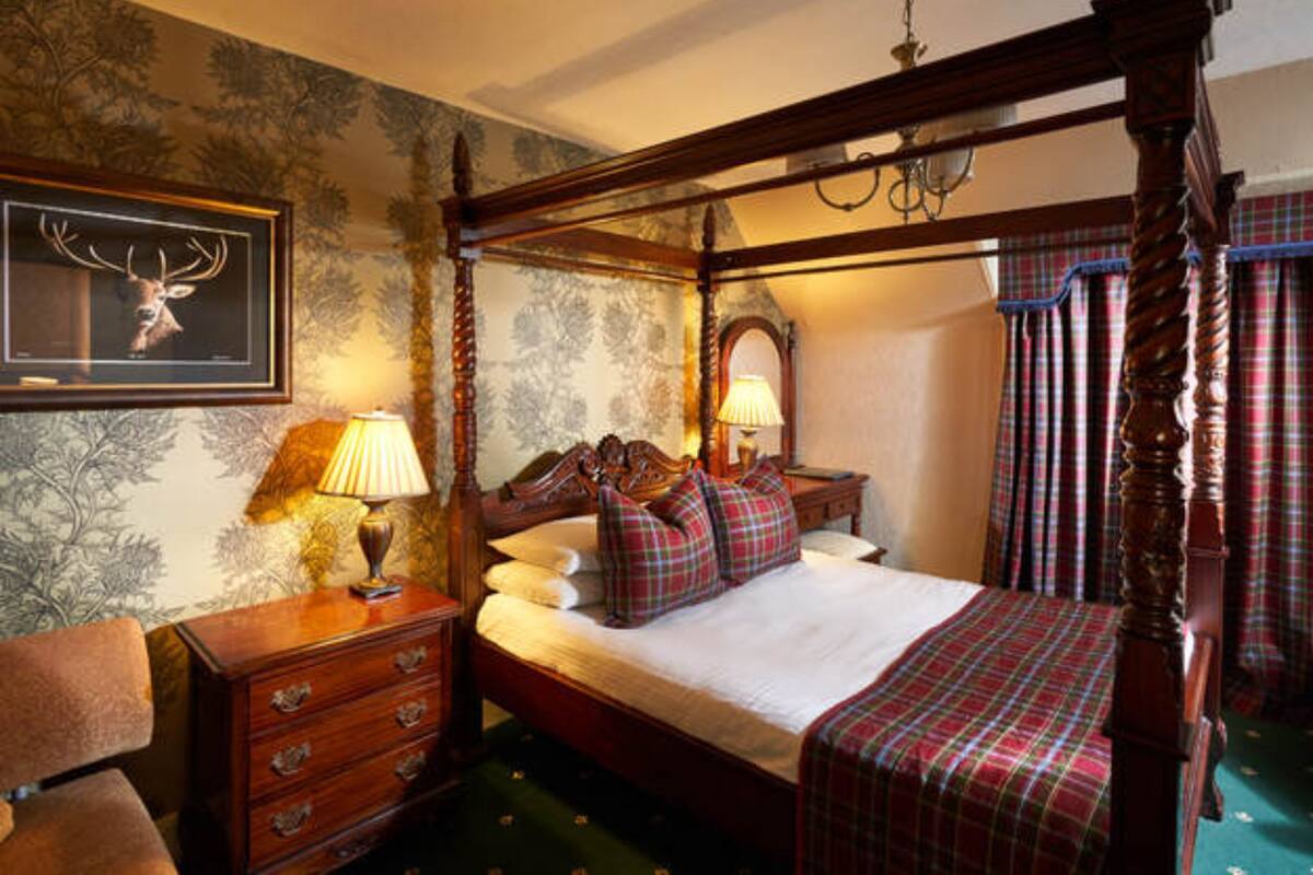 A four-poster bed with plaid bedding, a wooden dresser with drawers, a framed deer head on the wall, and a lamp on the dresser.