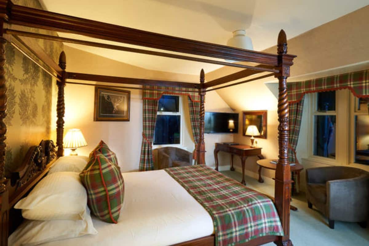 A four-poster bed with a plaid bedspread, a desk, and a chair in a room with a sloped ceiling and a window with curtains.