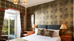 A bedroom with a bed, two bedside tables, a dresser, a desk, a chair, a balcony with a view, and a chandelier. - Loch Kinord Hotel (Aboyne)