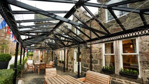 A covered outdoor seating area with wooden benches and tables, stone walls, and windows with curtains. - Loch Kinord Hotel (Aboyne)
