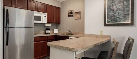A modern kitchen with a stainless steel refrigerator, a microwave, wooden cabinets, a granite countertop, and a small island with two chairs.
