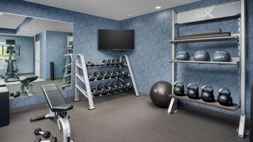 Un gimnasio con una banca, mancuernas, kettlebells y un televisor en la pared.