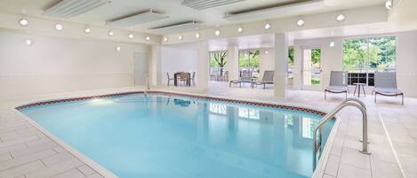 An indoor swimming pool with a tiled floor, a poolside seating area, and a view of the outdoors.