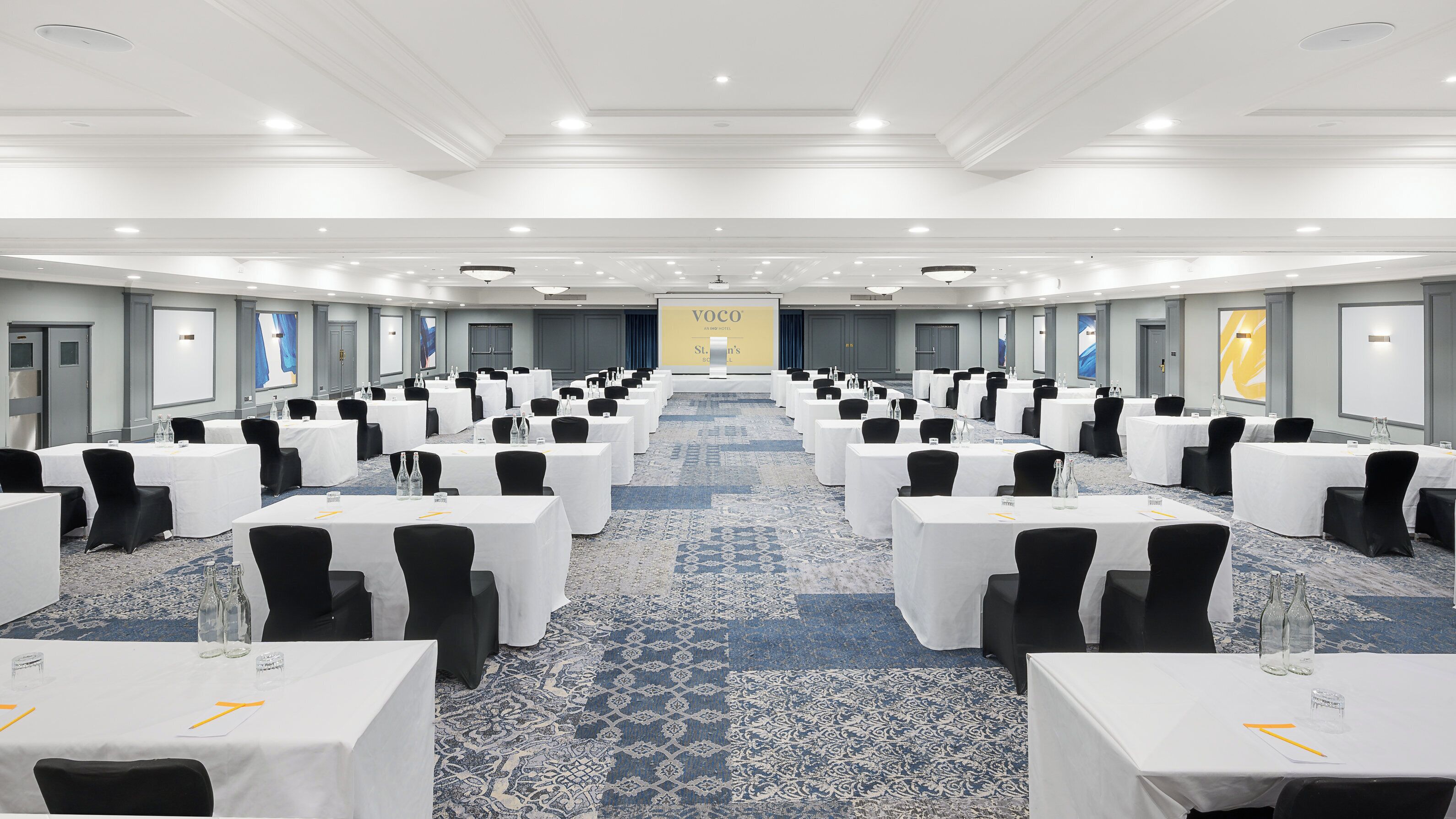 A conference room with rows of tables and chairs, a carpeted floor, and a large screen at the front.
