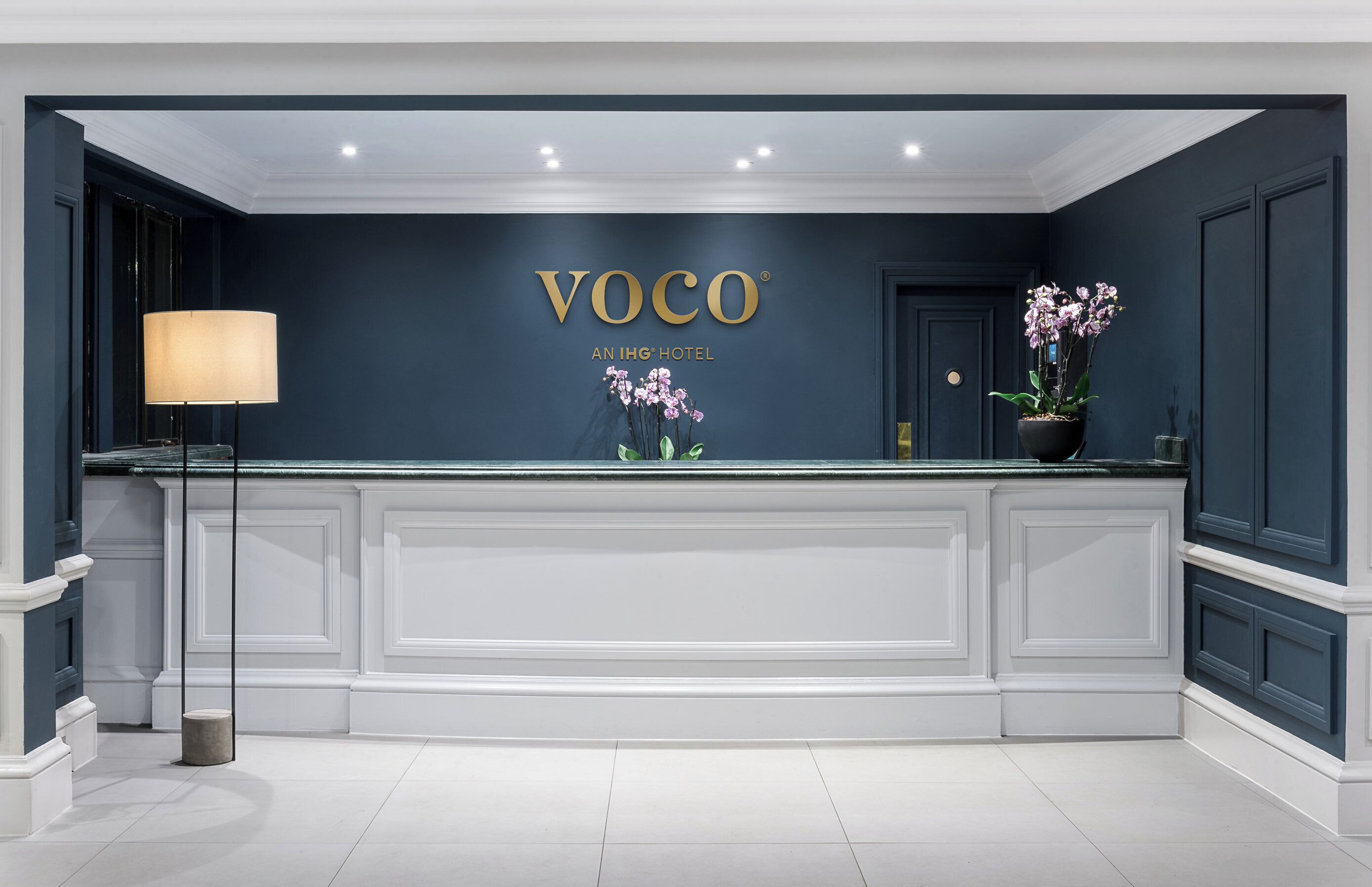A reception desk with a dark blue wall, a white marble counter, and a sign that reads 'VOCO AN IHG HOTEL'.