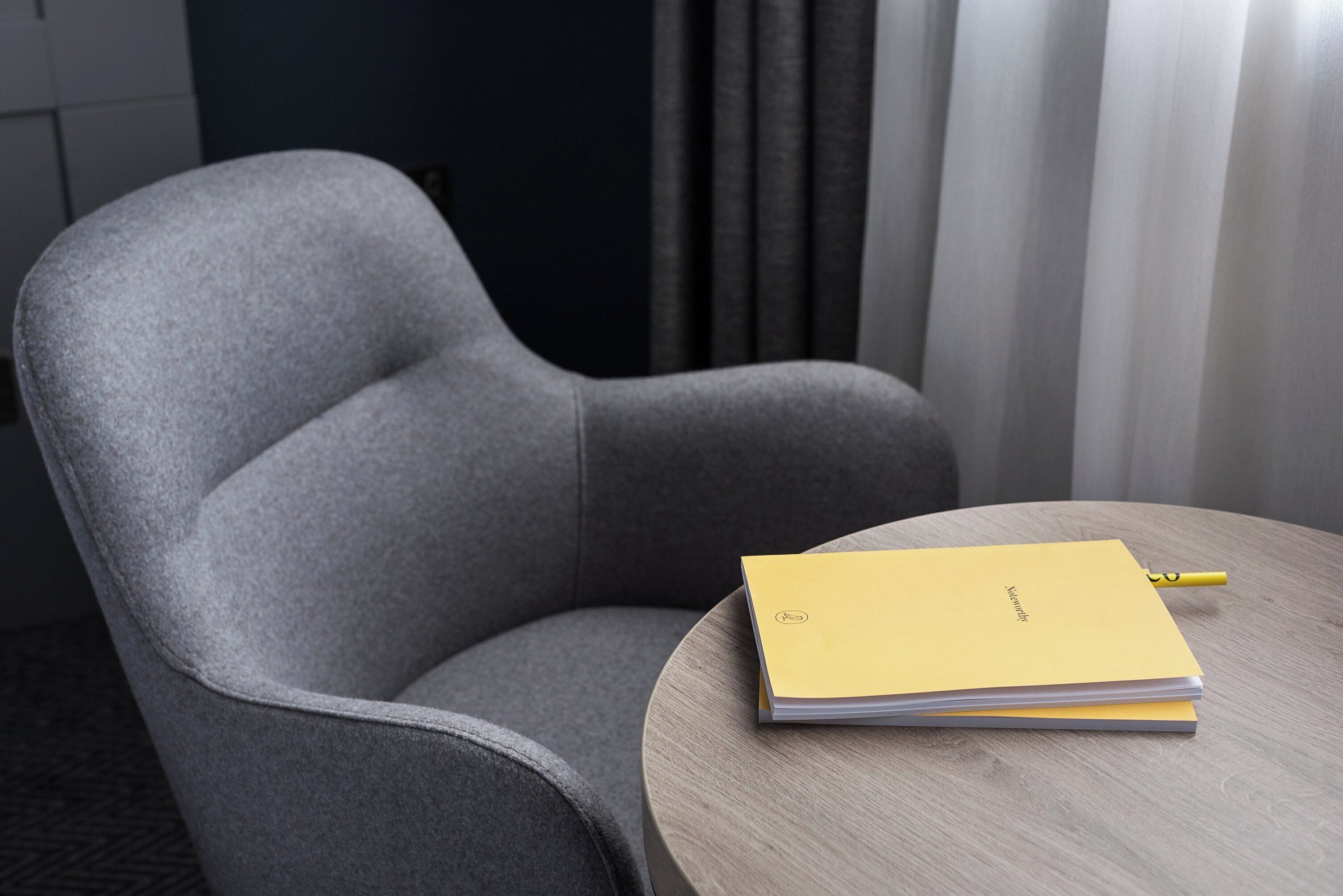 A grey upholstered armchair with a wooden table beside it, holding a yellow folder and a pen.