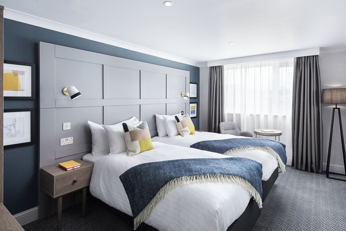 A hotel room with two beds, a large headboard, a bedside table with a lamp, a small round table, and a floor lamp.