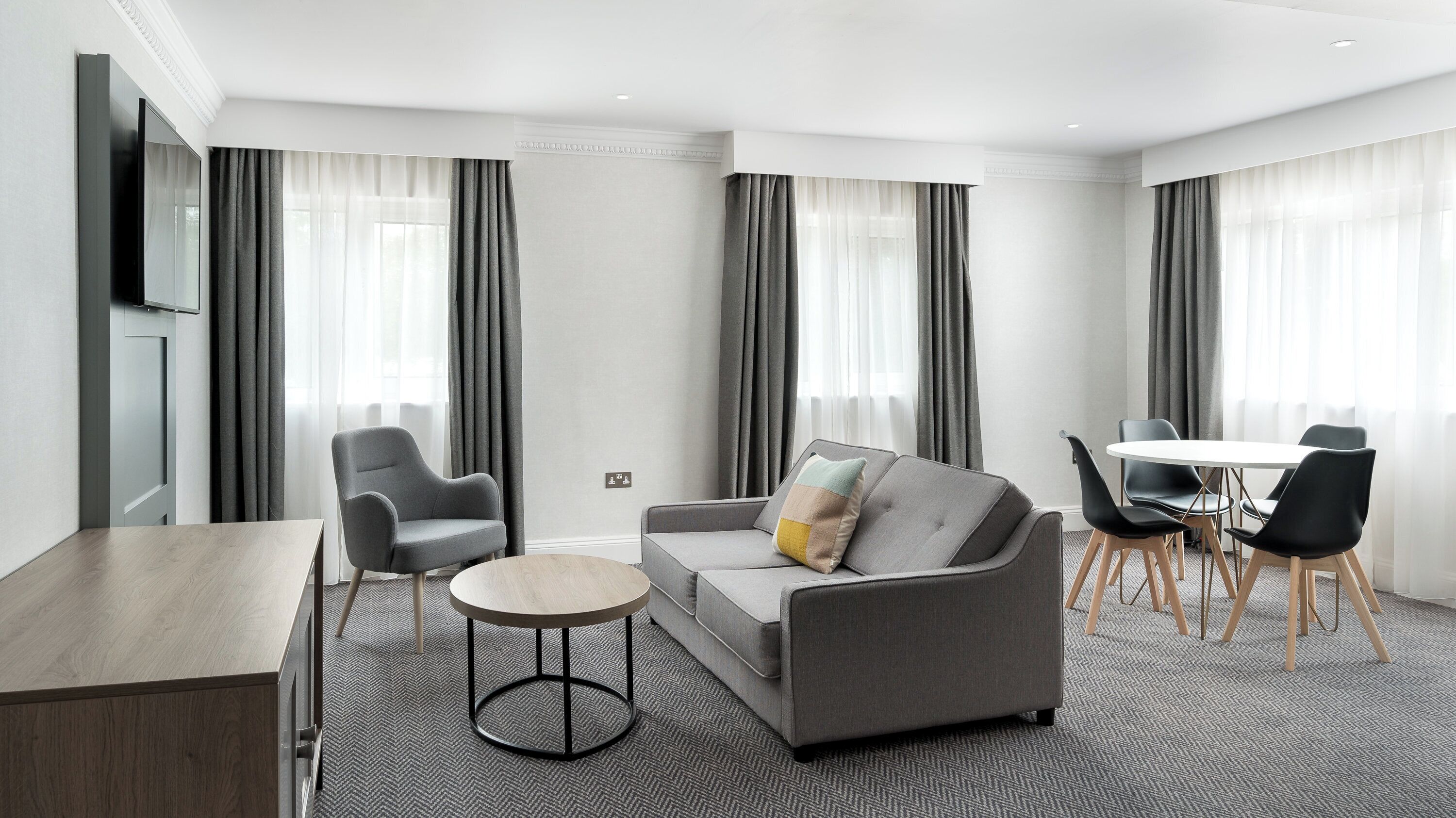 Een moderne hotelkamer met een grijze bank, een ronde salontafel, een eettafel met stoelen en grote ramen met grijze gordijnen.