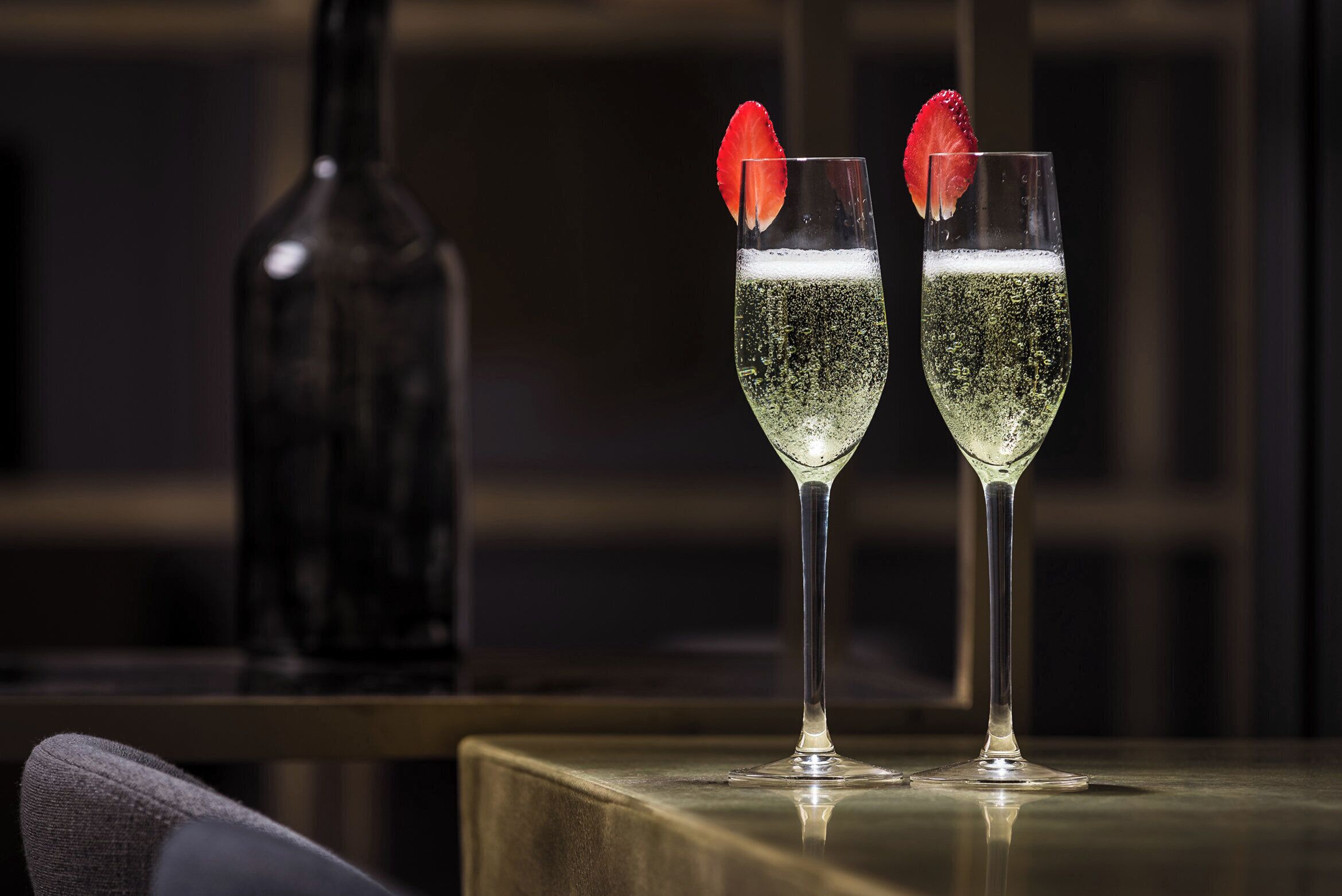 To glas champagne med jordbær på kanten, placeret på en reflekterende overflade med en flaske i baggrunden.