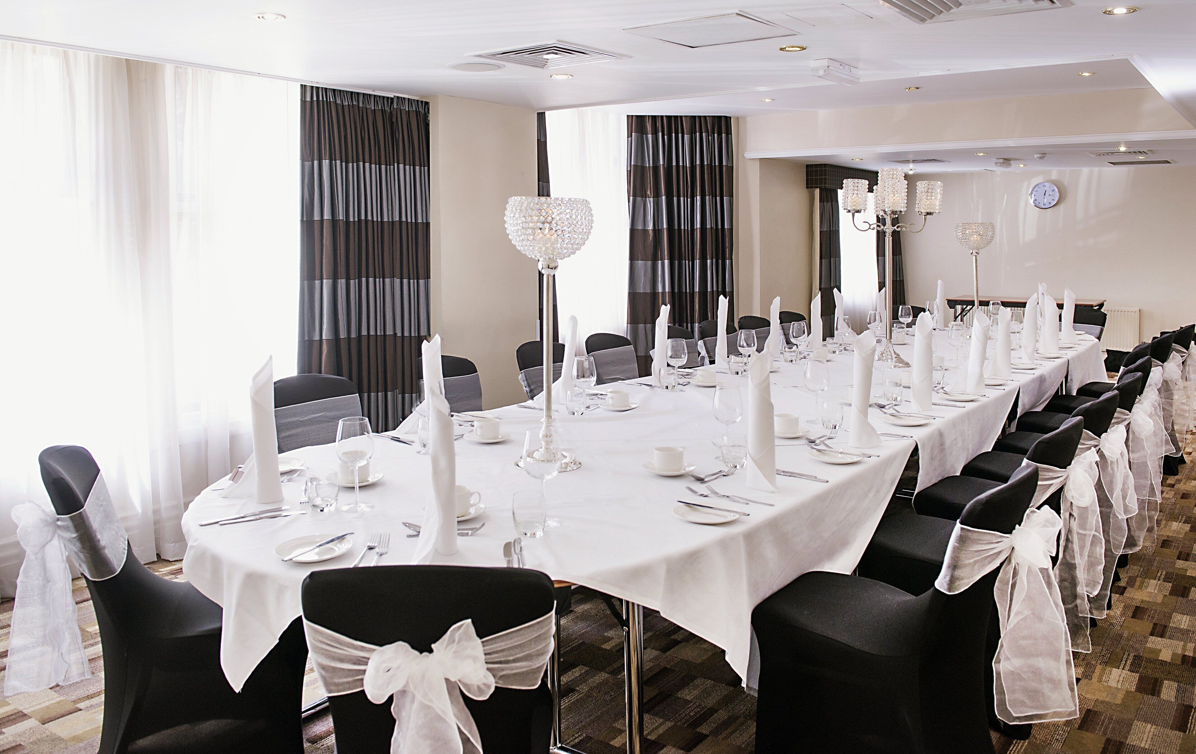 Una larga mesa banquete dispuesta con manteles blancos, sillas negras con cintas blancas y candelabros de cristal.