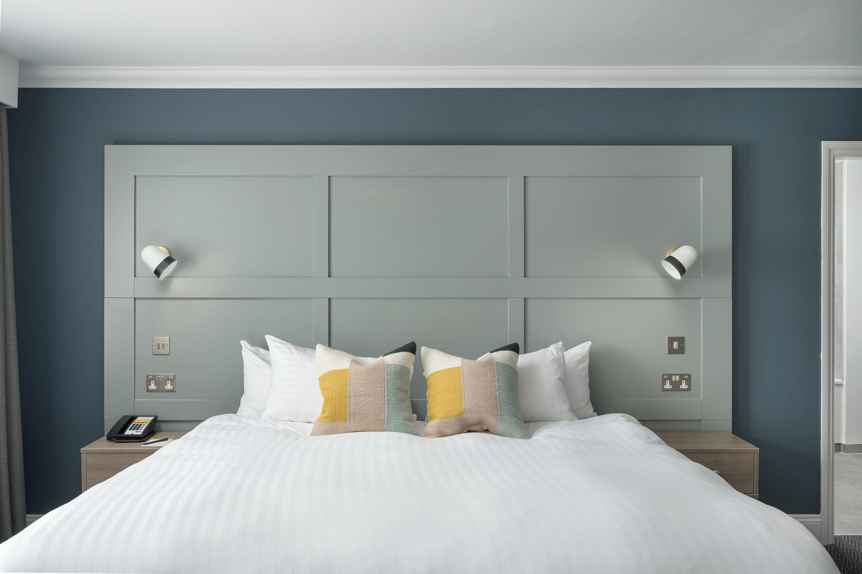 A neatly made bed with a white comforter and decorative pillows, a bedside table with a phone, and a wall-mounted light fixture.